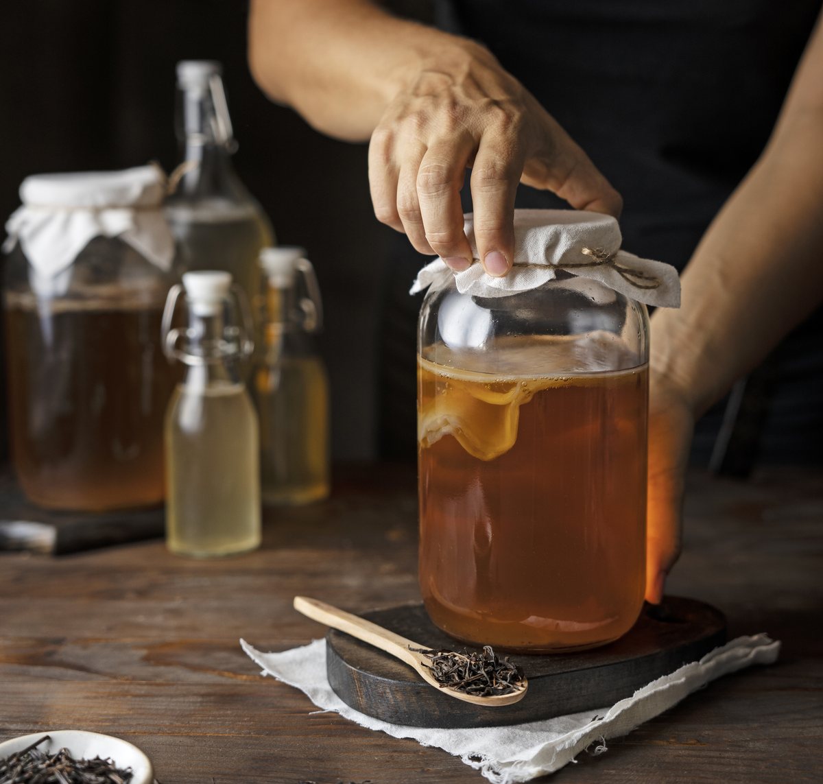A maker lifting the cloth cover from a jar of kombucha resting on a workbench