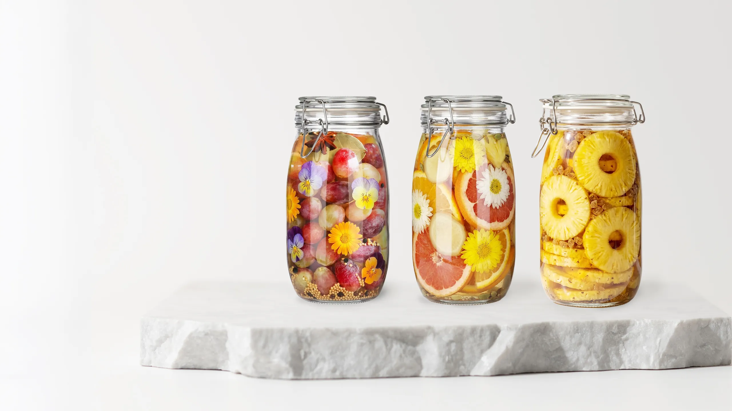 Three glass jars of fruit and flowers fermenting, on a marble slab