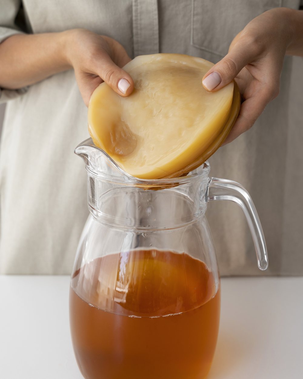 Hands lifting a SCOBY culture from a jug of brewing kombucha